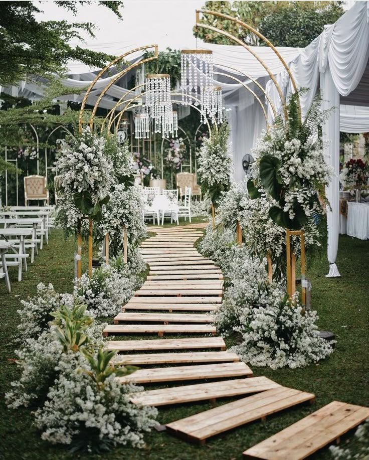 Outdoor wedding ceremony aisle with a wooden path, gold metal arches, and white floral arrangements.